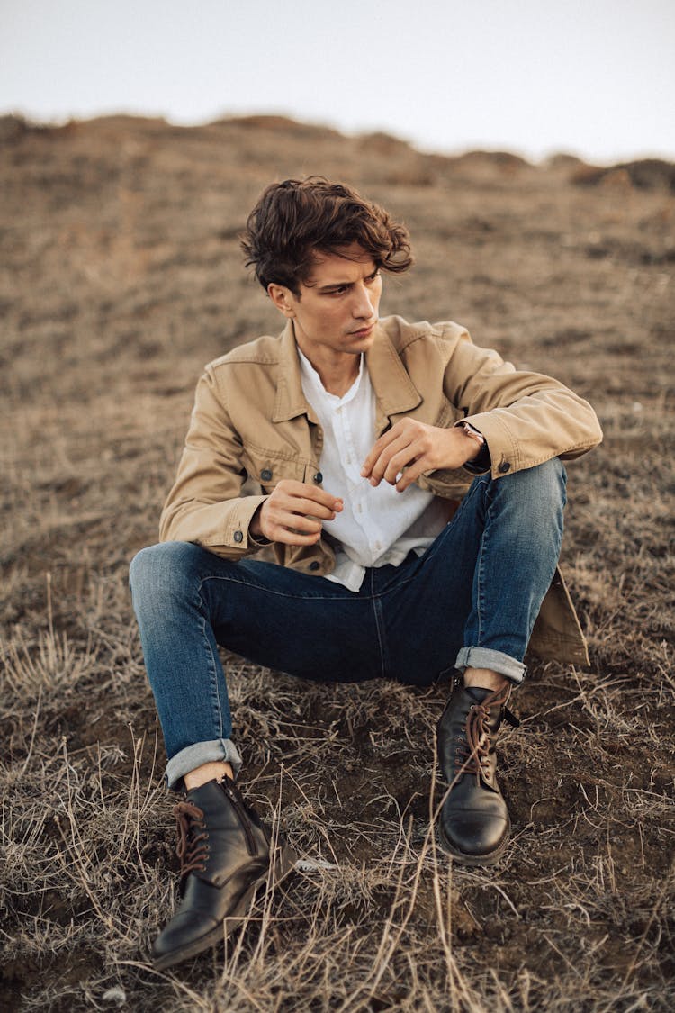 Young Man In Blue Jeans And Beige Jacket Sitting On Ground In Empty Fields