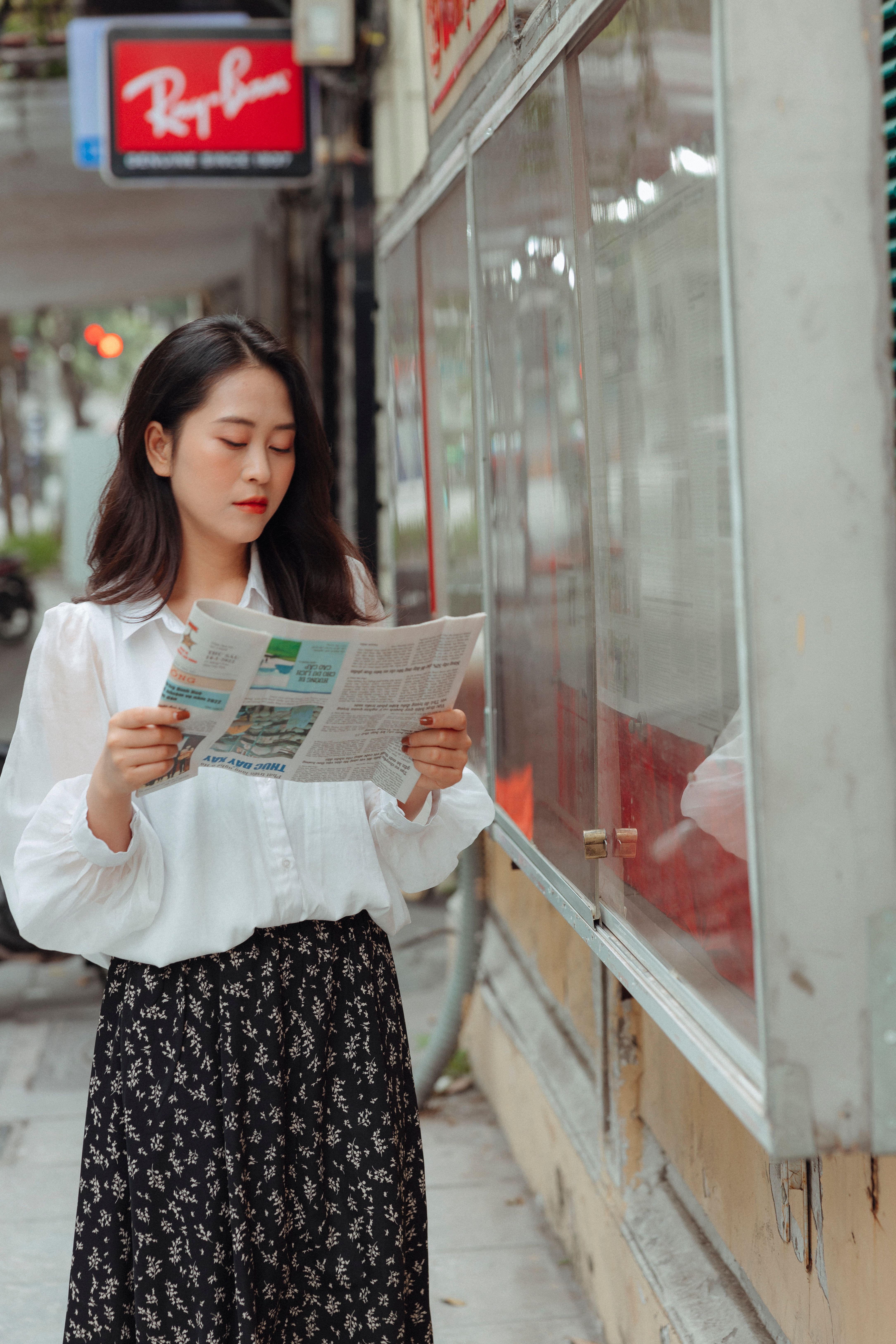 Fashionable woman in white blouse reads newspaper while standing on city street.