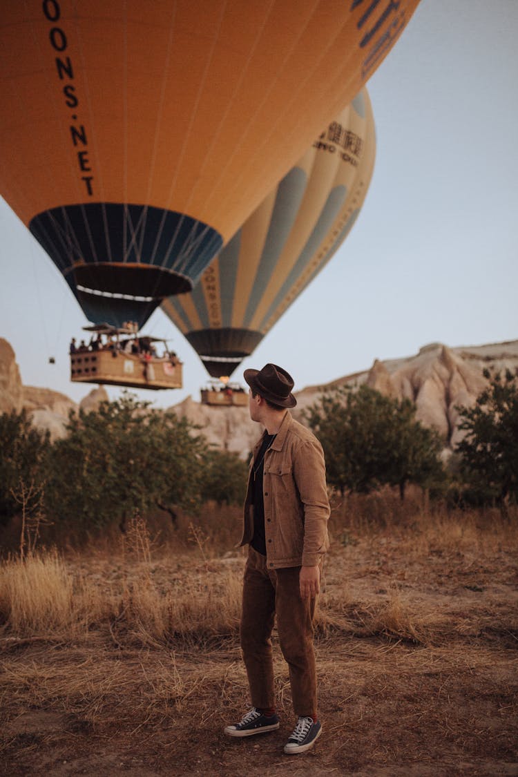 Man In Hat Standing In Fields And Looking Back At Balloons Hovering Over Ground