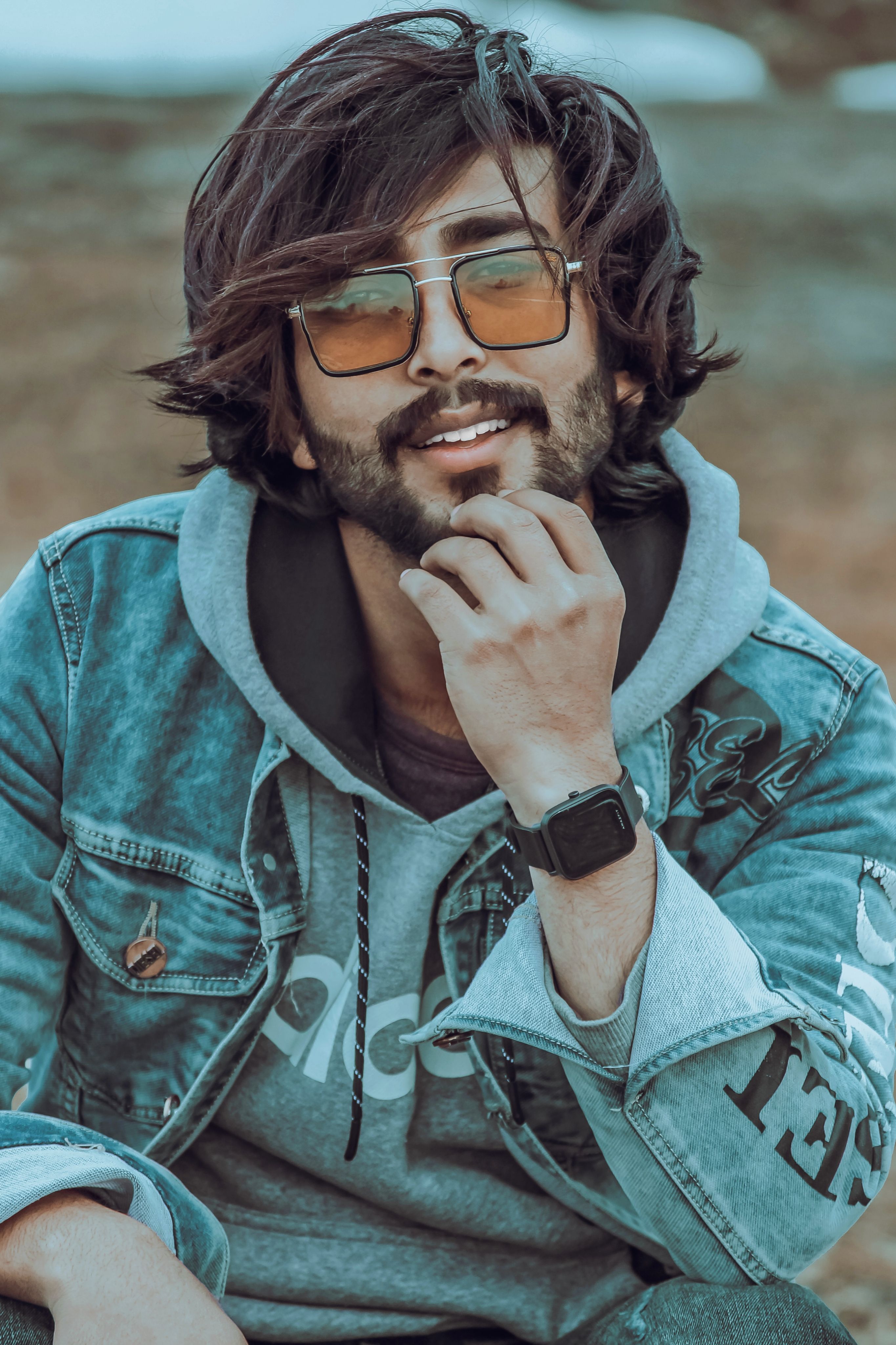 Young man with sunglasses and beard, smiling outdoors in casual denim and hoodie.