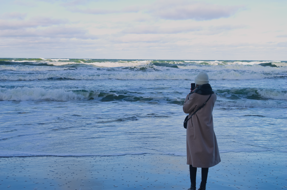 Person in winter attire capturing ocean waves at the beach.