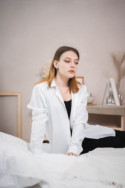 Free A contemplative young woman sits on a bed in a white shirt, creating a serene and stylish vibe. Stock Photo