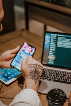 Close-up of a tattooed hand operating a smartphone and laptop on a wooden table indoors.