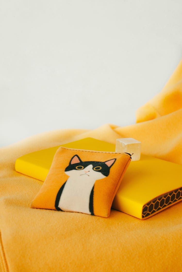 A Cute Cat Logo On A Coin Purse Beside A Book On A Yellow Surface