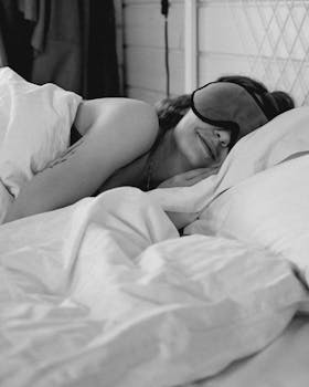 Black and white photo of a woman smiling in bed with an eye mask.