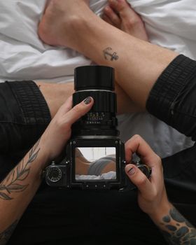 Close-up view of tattooed hands holding a camera, capturing a reflective image on a bed scene.