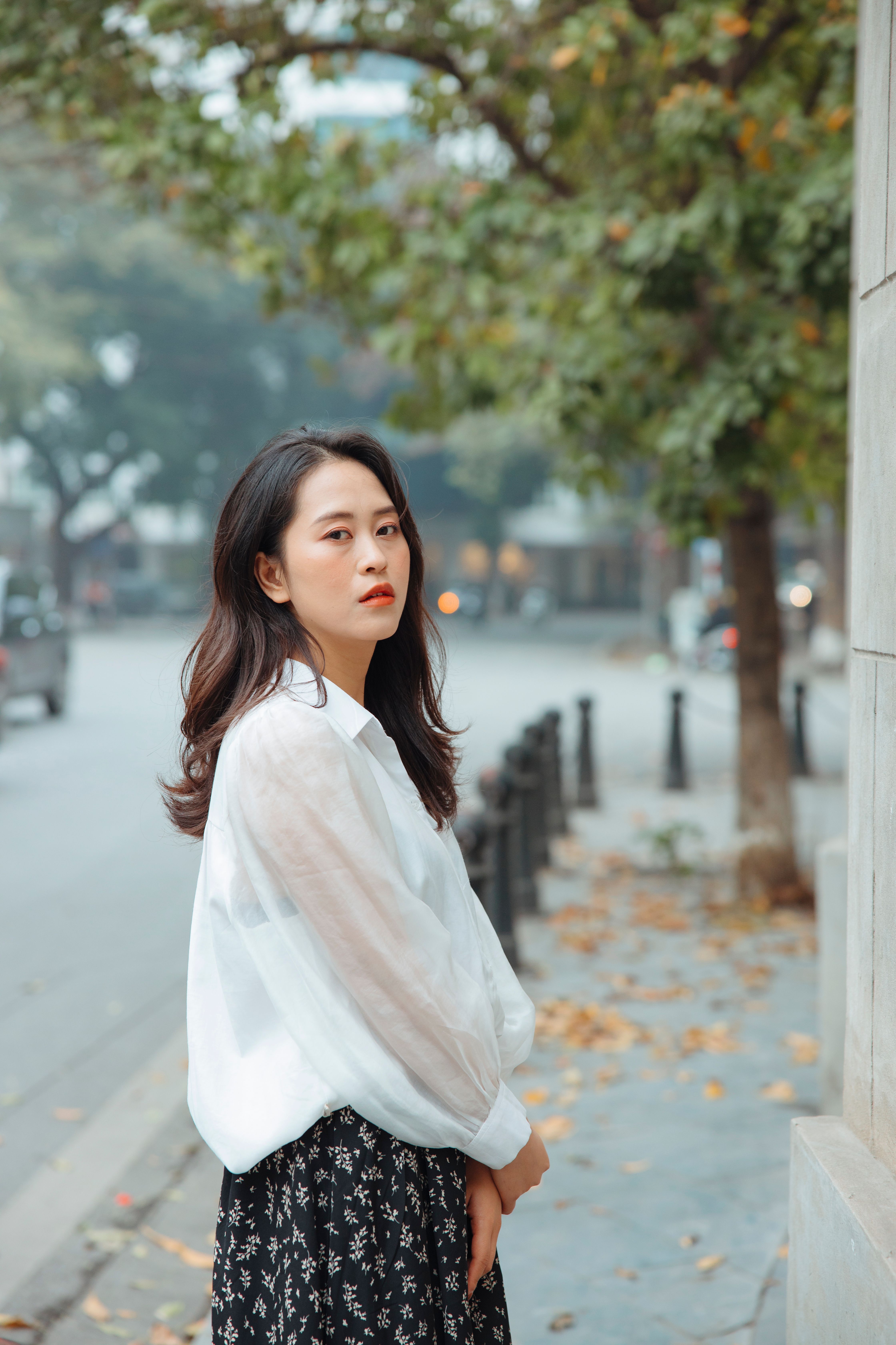 Stylish Asian woman in a white blouse stands elegantly on a tree-lined street.