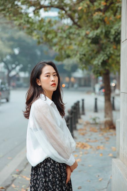 Stylish Asian woman in a white blouse stands elegantly on a tree-lined street.