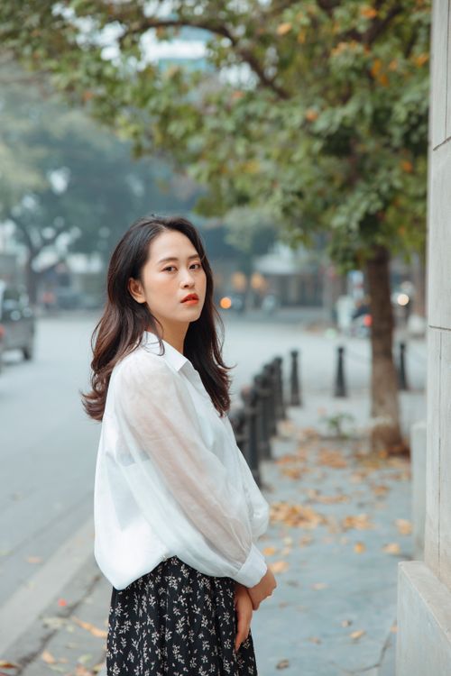 Free Stylish Asian woman in a white blouse stands elegantly on a tree-lined street. Stock Photo