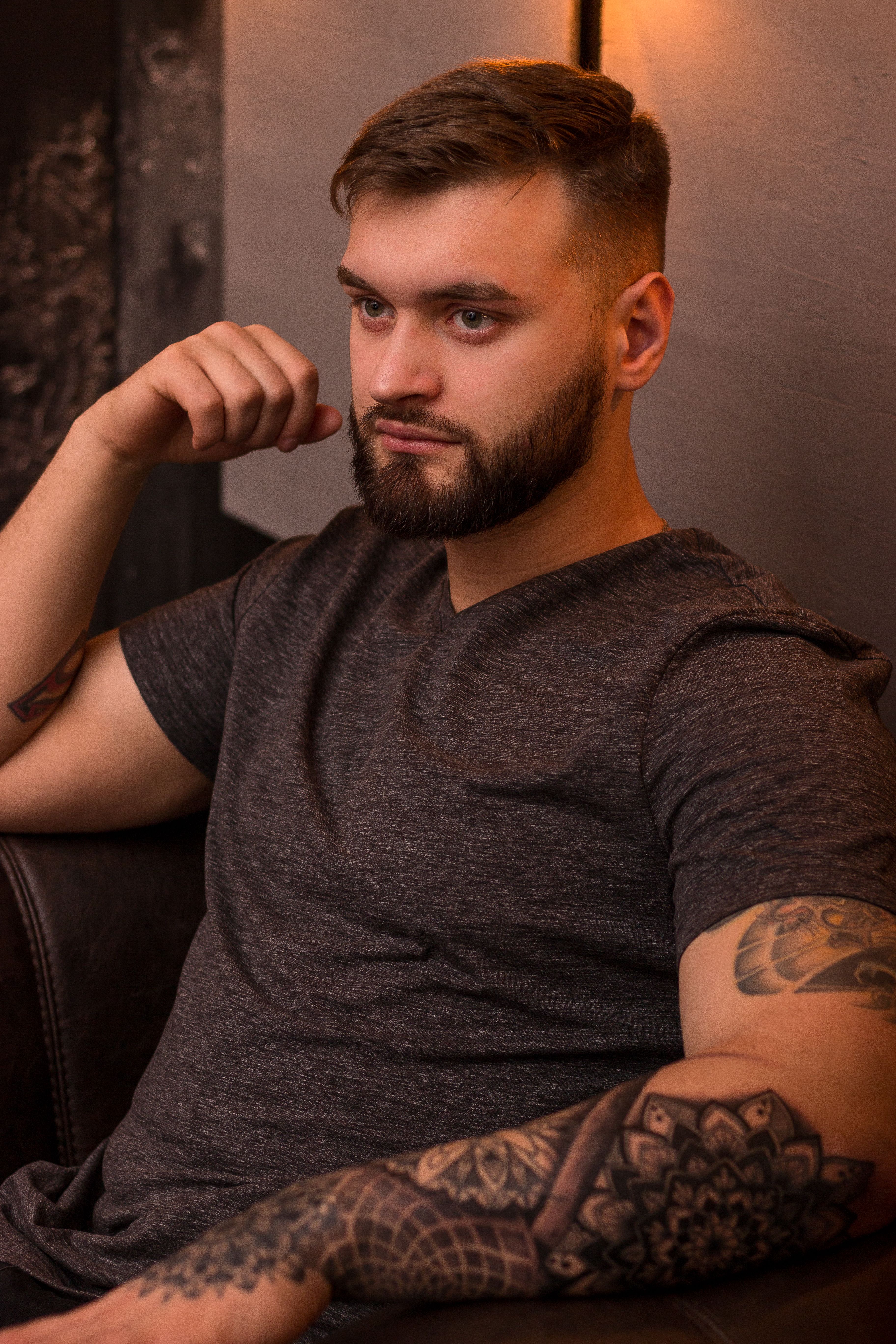 Portrait of a stylish bearded man with tattoos, wearing a gray shirt, sitting and posing indoors.