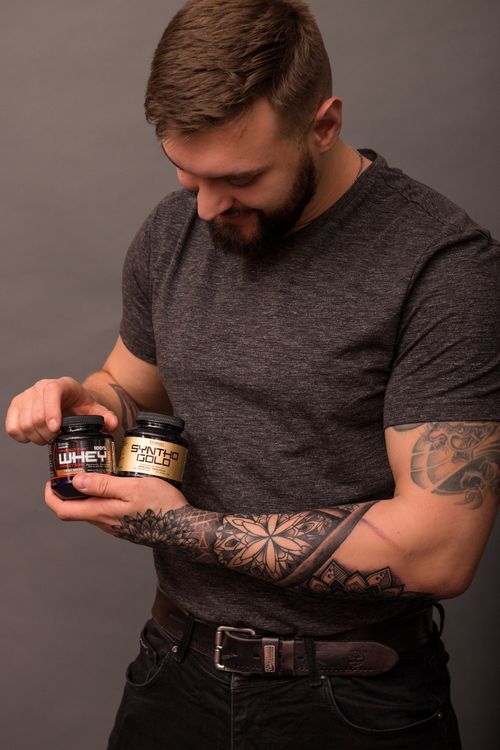 Free Bearded man with arm tattoos holding protein supplement bottles in a studio setting. Stock Photo
