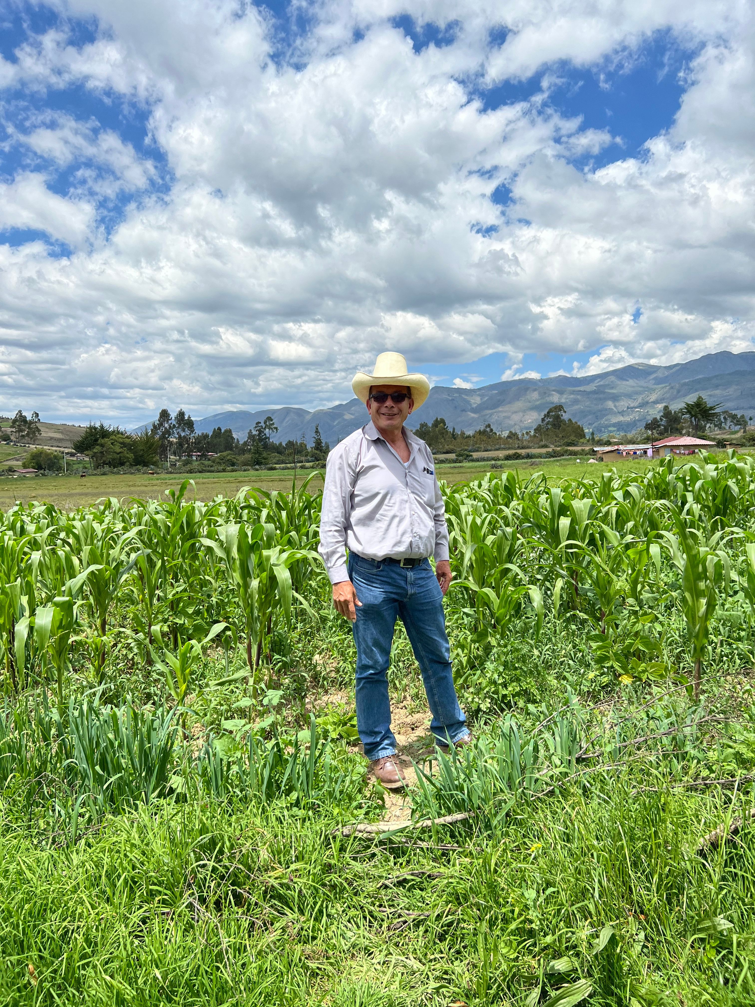 A senior farmer stands proudly in a lush cornfield under a bright, cloudy sky, showcasing rural agriculture.