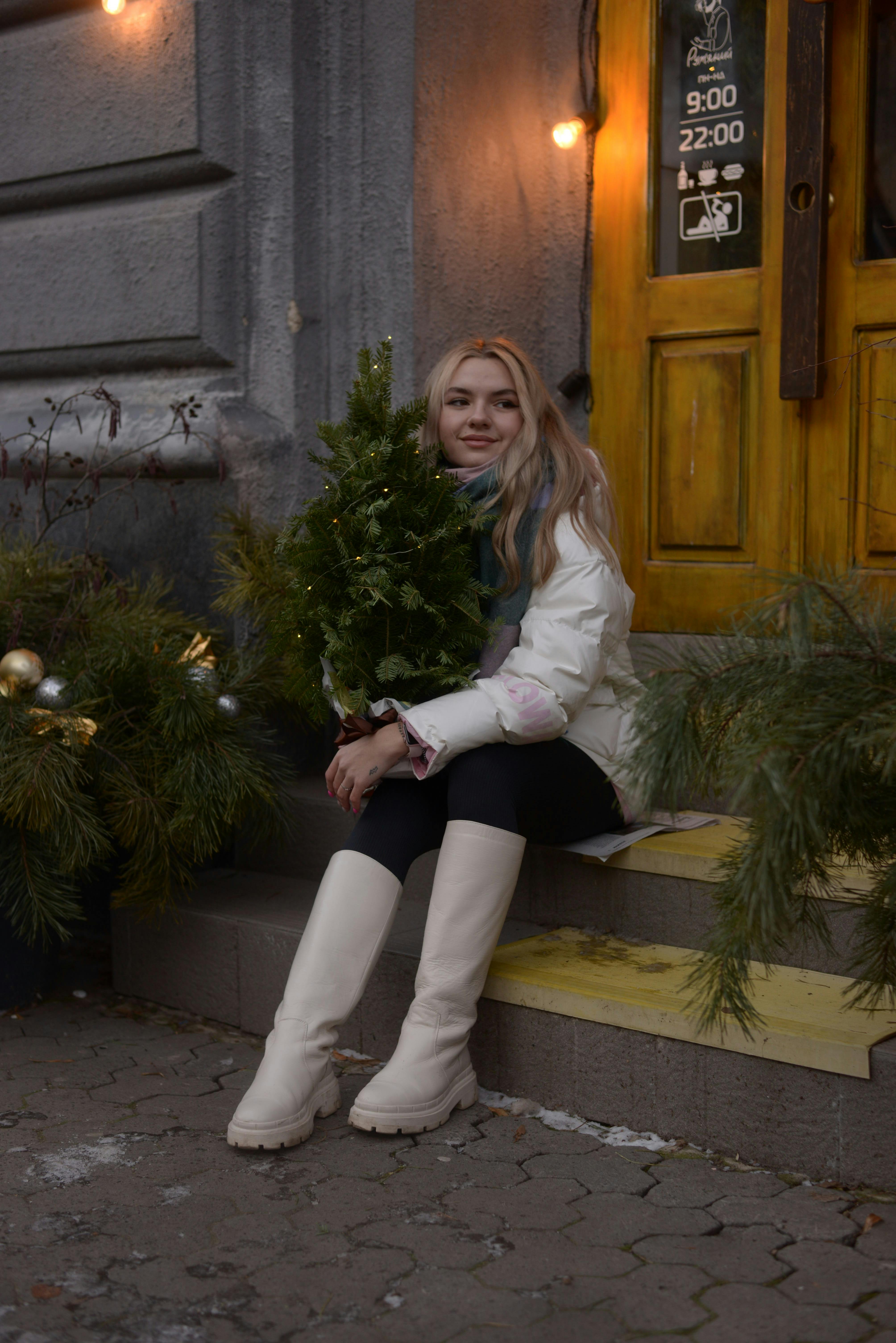 A woman sits on steps holding a small Christmas tree outside a decorated building in winter.