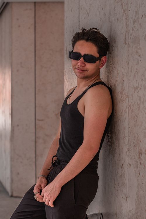 Free Man in sunglasses poses confidently against a textured wall, wearing a black tank top. Stock Photo