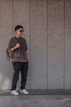 Young man in sunglasses posing against wall outdoors, casual style.