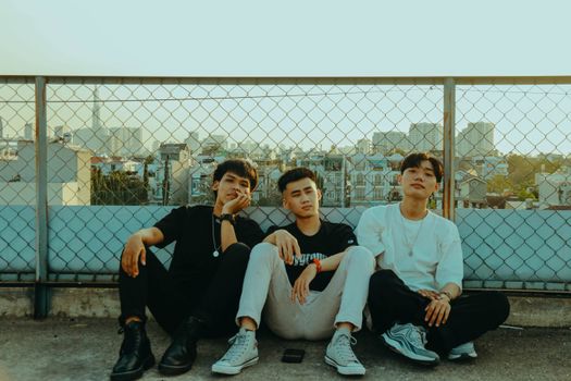 Three stylish men sitting against a fence with a cityscape in the background, enjoying the sunny day.