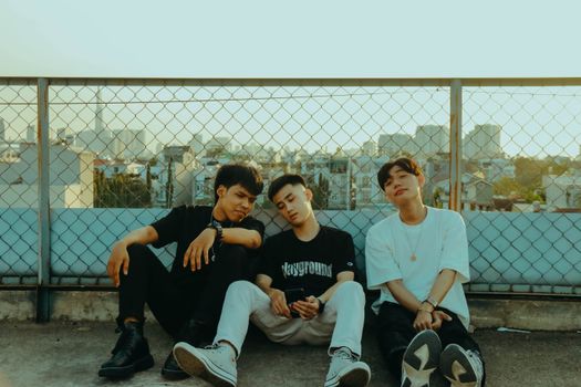 Three young men relaxing on a city rooftop with a metal fence and skyline backdrop.