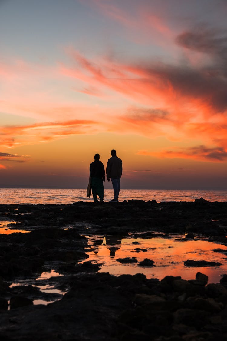 Silhouettes Of Two Unrecognizable People Standing At Seashore And Admiring Colourful Sunset