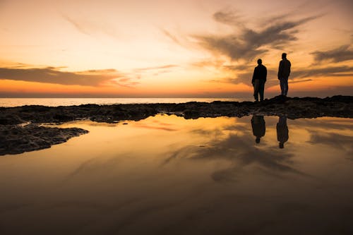 Silhouette of Two People Standing on the Rocks Near a Body of Water