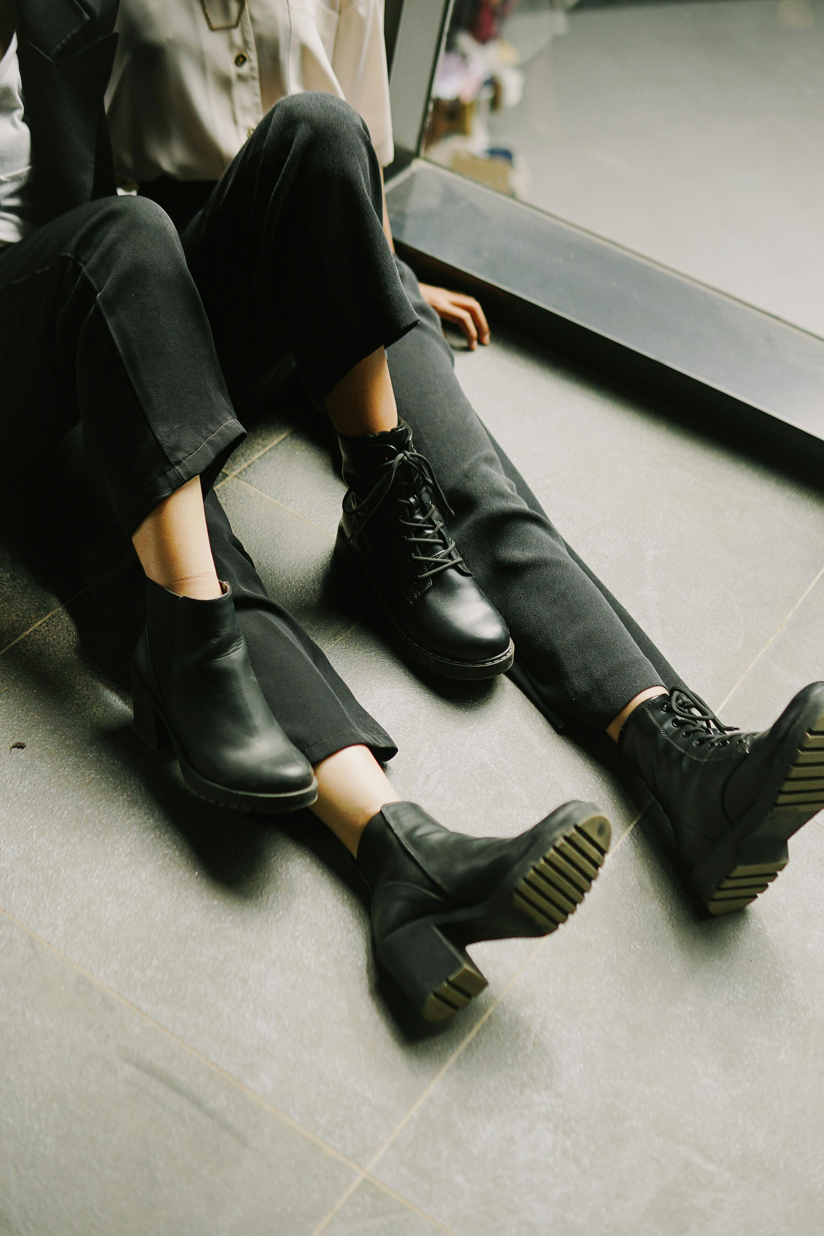 Two individuals in stylish black boots sitting indoors on a tiled floor.
