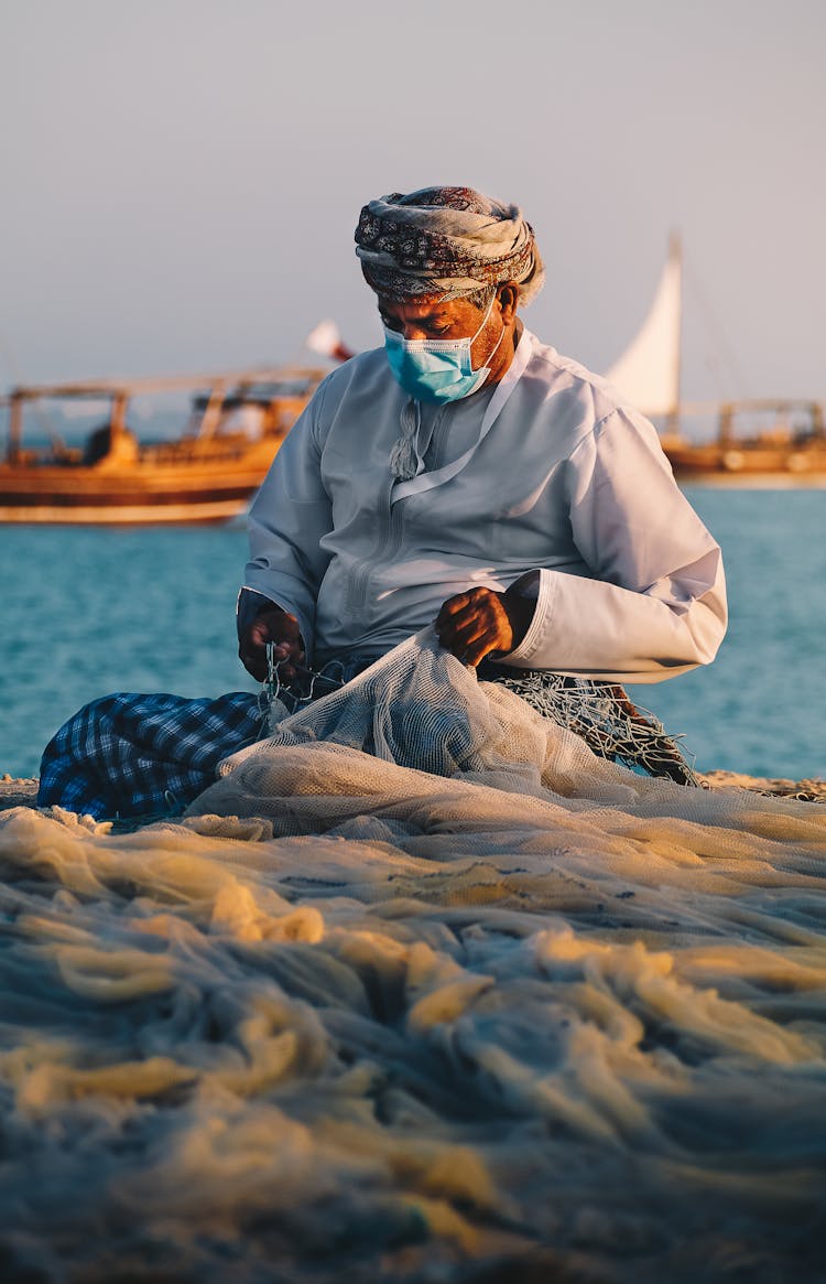 Fisherman Sitting At Seashore And Mending Fish Net