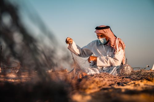 Free A Middle Eastern fisherman wearing a mask mends a fishing net in Doha, Qatar, against a warm sunset backdrop. Stock Photo