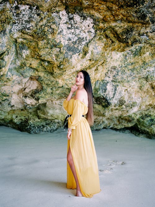 Free A woman in a yellow dress poses by rocky formations on a sandy Bali beach. Stock Photo