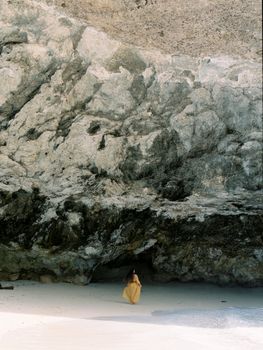 Woman in yellow dress stands by rocky beach cave in Bali's serene landscape.