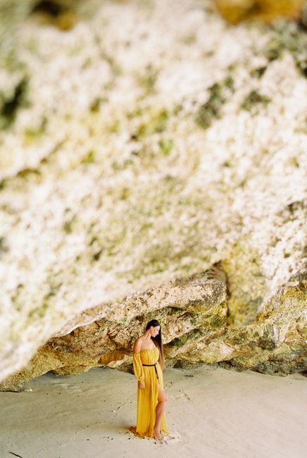 Woman in yellow dress standing under rock formation on Bali beach, evoking a serene and scenic atmosphere.