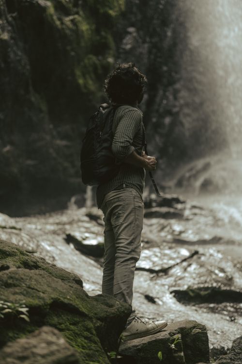 Free A man stands facing a misty waterfall in a lush, rocky forest setting. Stock Photo