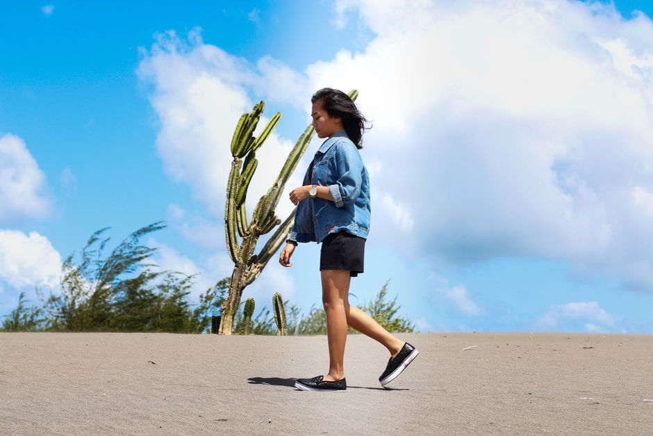 A woman in casual wear walks through a sandy desert under a bright blue sky with cacti.