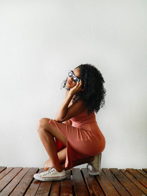 Fashionable young woman with curly hair squatting on wooden floor, wearing sunglasses and a dress.