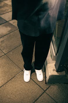 A person wearing white sneakers standing on a city sidewalk at night.
