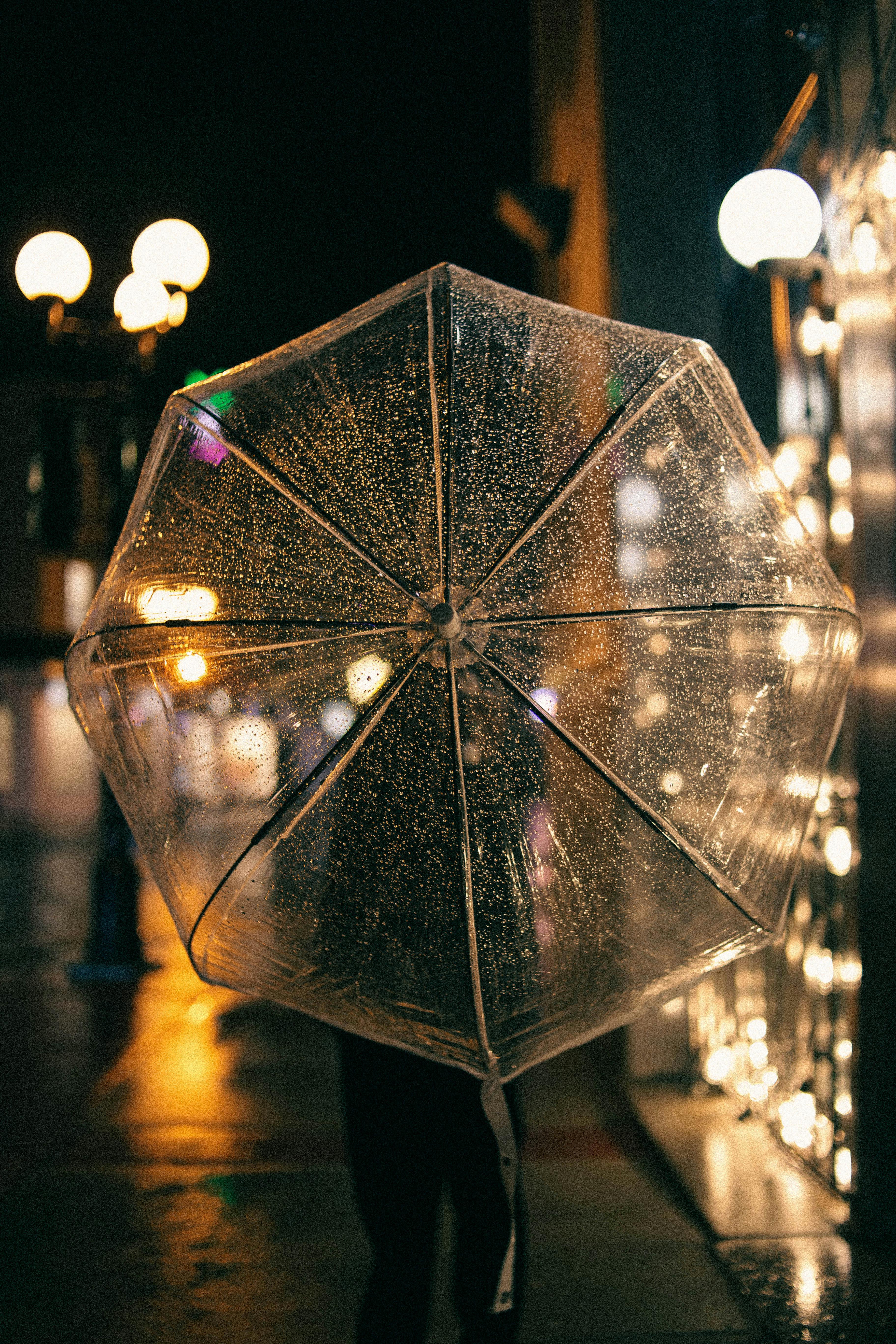 A transparent umbrella capturing raindrops in a warmly lit urban street at night.