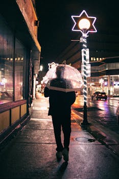 A person walks with an umbrella on a rainy night city street, illuminated by streetlights.