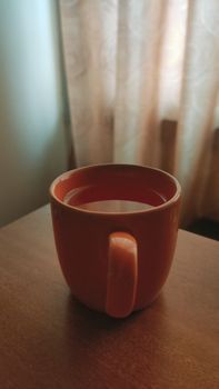 A cozy close-up of a ceramic mug with coffee placed on a wooden surface by the window.
