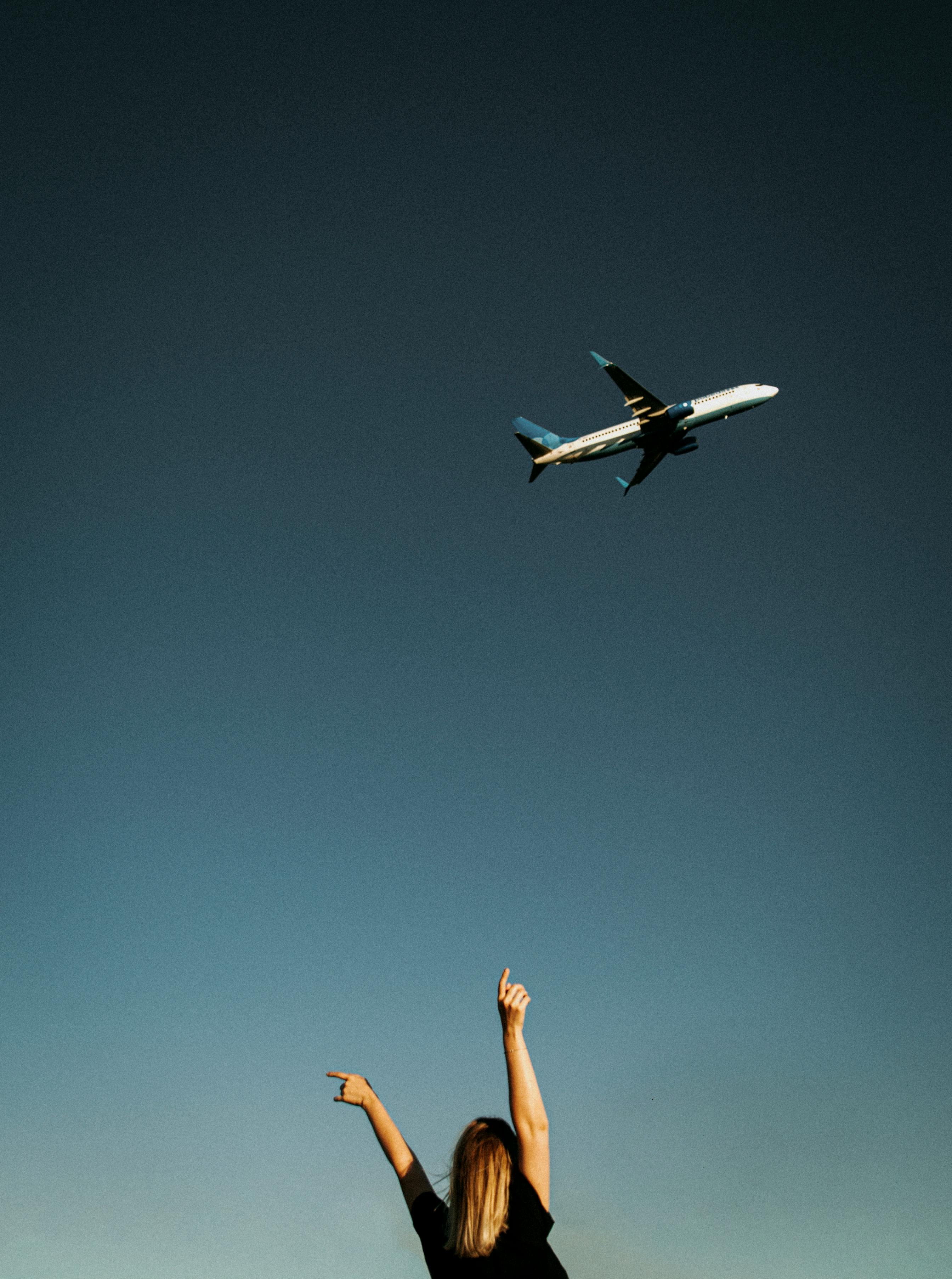A woman points at a flying airplane against a clear blue sky, symbolizing travel and freedom.