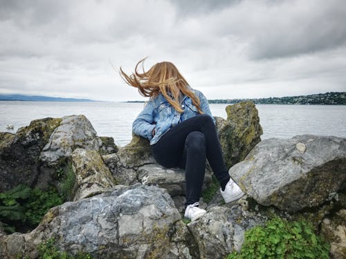 無料 青いデニムの襟付きボタンアップジャケットと灰色の岩の上に座ってレイクビューを見ている黒のジーンズを持つ女性 写真素材
