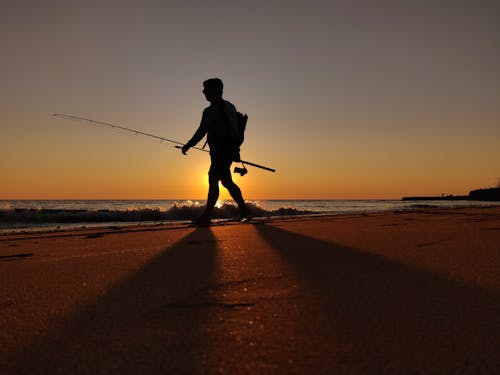 Silhouette of a Man Carrying a Fishing Rod while Walking on the Sea Shore during Sunset