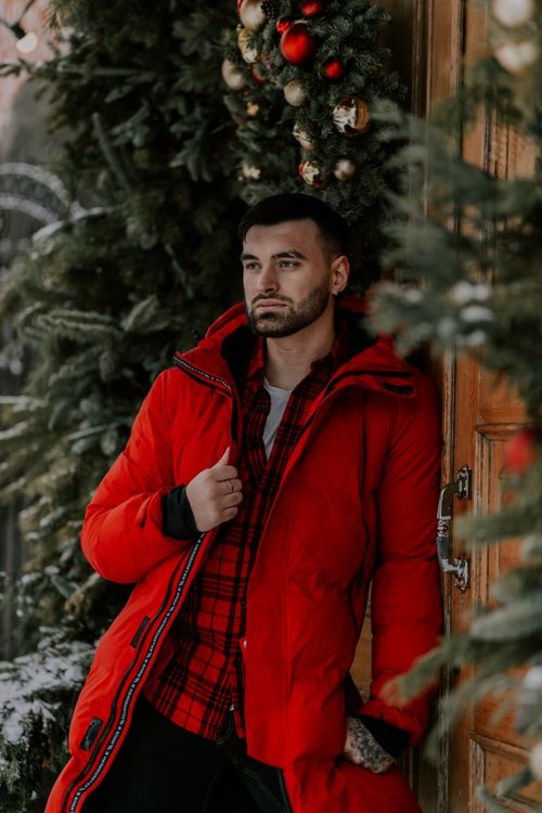 Free Man leaning against a door wearing a red coat with Christmas wreaths, conveying a winter holiday vibe. Stock Photo