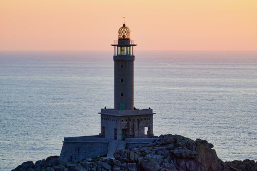 Free A solitary lighthouse stands against an orange sky over a tranquil sea, showcasing serene coastal beauty. Stock Photo
