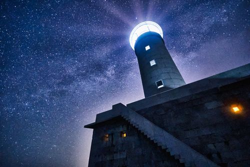 Free Low-angle view of a lighthouse illuminated against a starry night sky, creating a dramatic celestial scene. Stock Photo