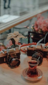 Two vintage cameras and a glass of tea on a wooden table with flowers in the background.