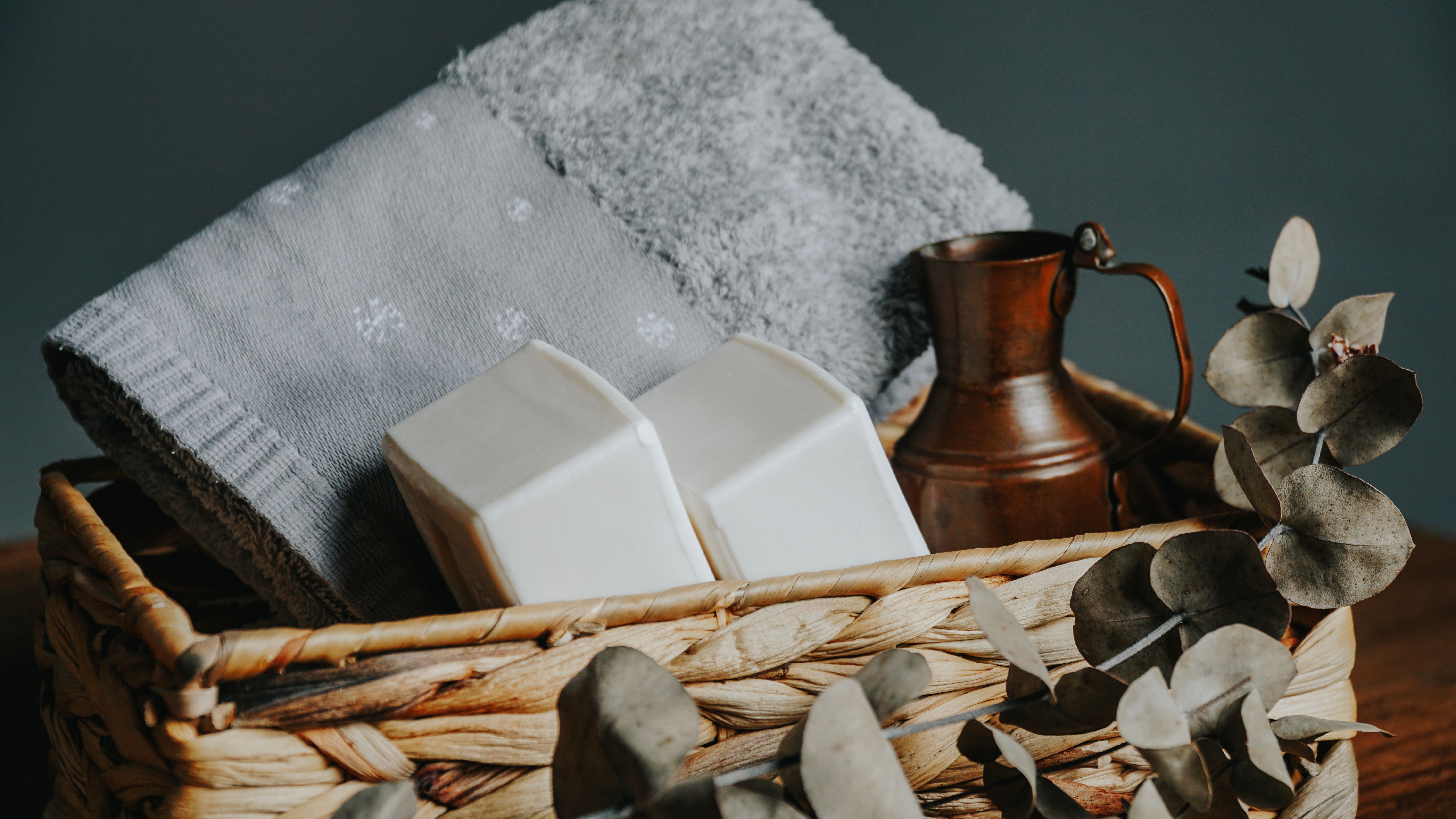 A woven basket holds towels, soap, a copper pitcher, and eucalyptus leaves, creating a calming spa atmosphere.