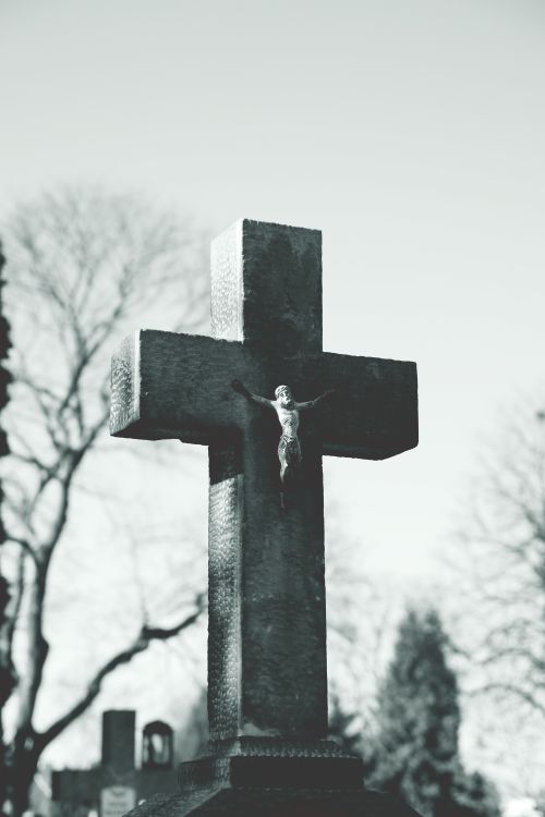 Free Monochrome photo of a cemetery cross with Jesus Christ in a serene outdoor setting. Stock Photo