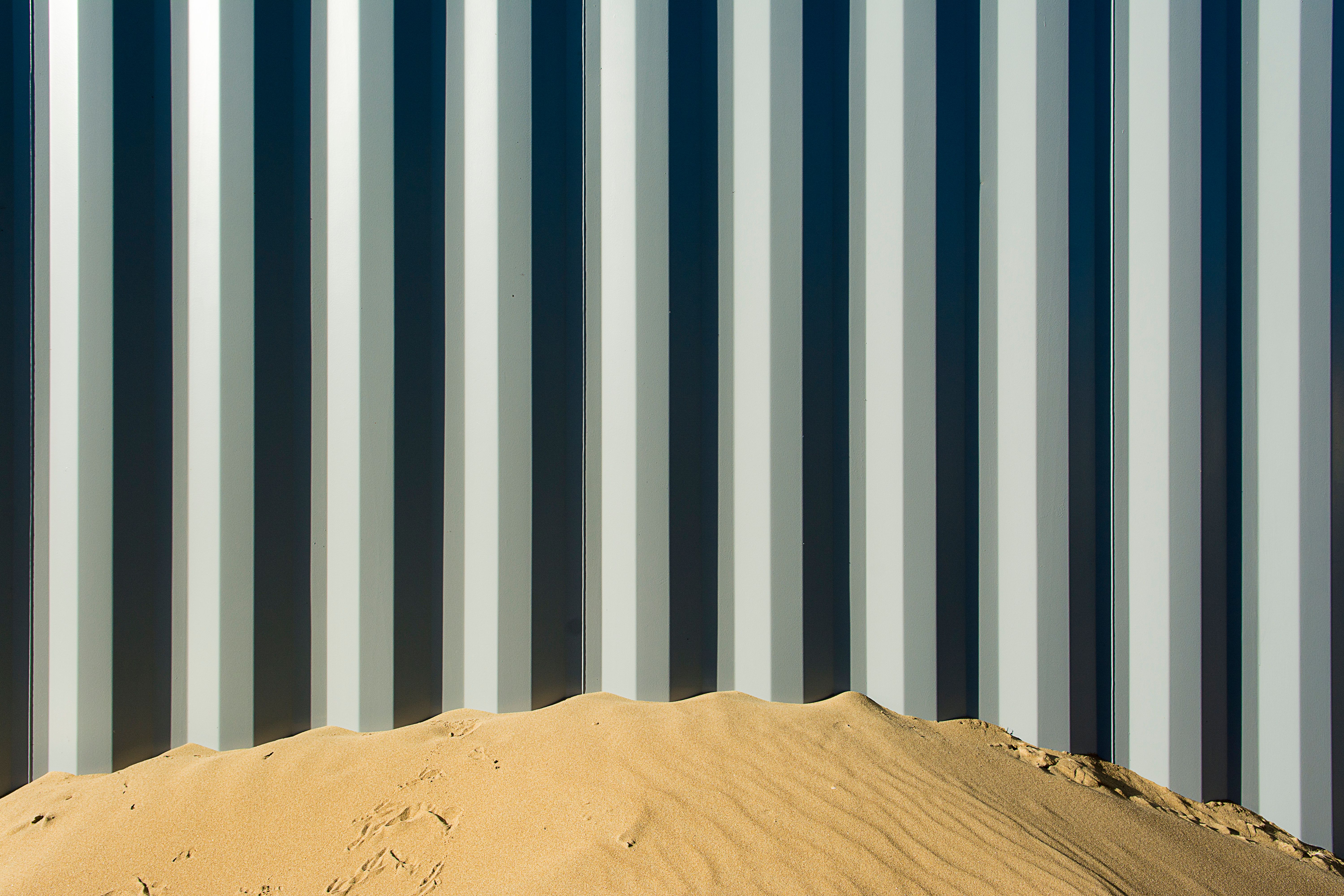 A corrugated metal fence contrasts against a small sand dune under bright lighting.