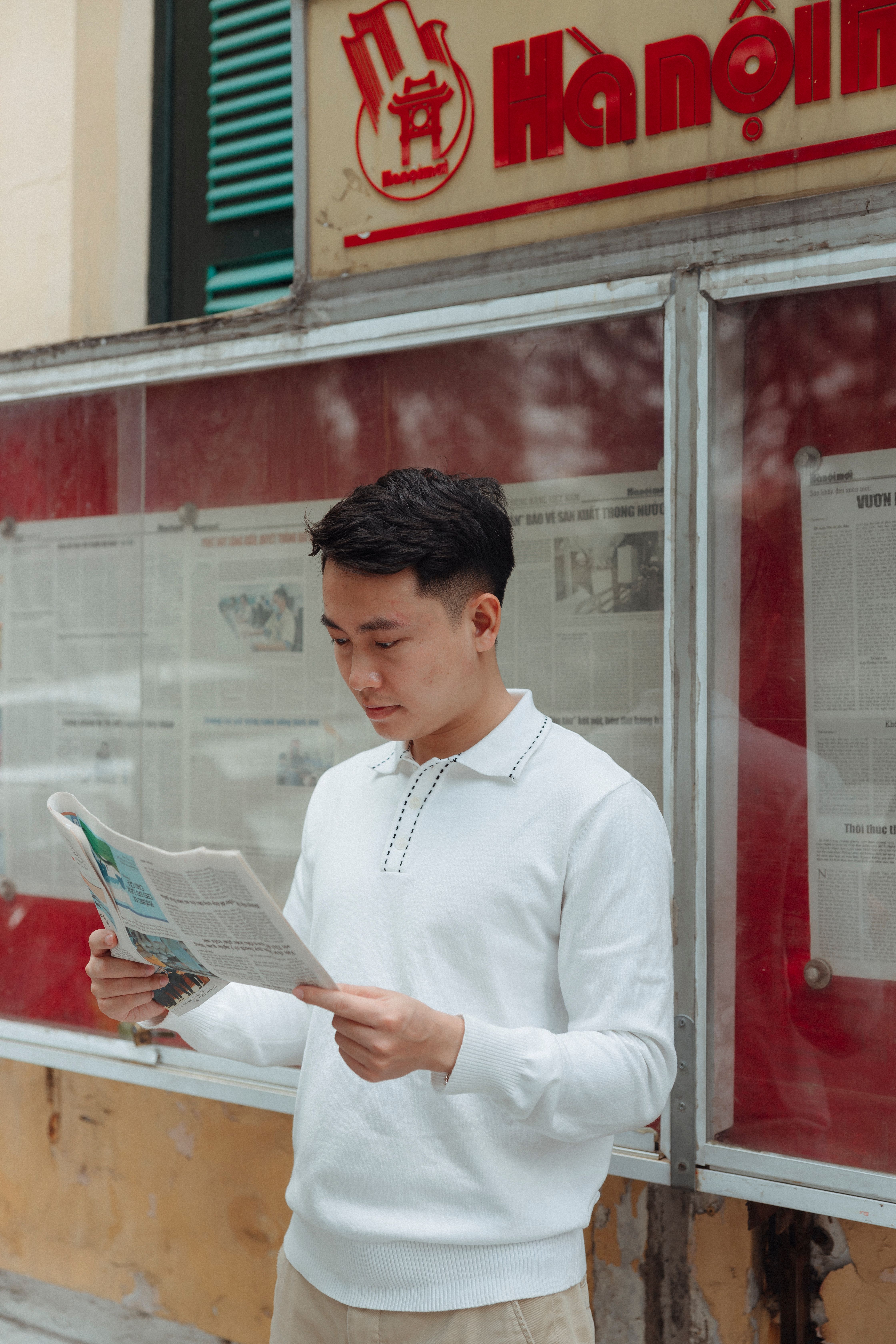 Asian man reading a newspaper outdoors in Hà Nội, Vietnam near Ha Noi Moi building.