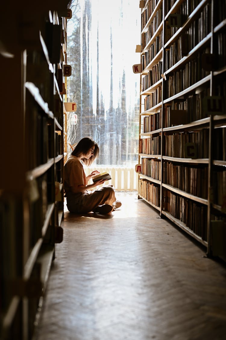 Girl Sitting On Library Floor Reading A Book 