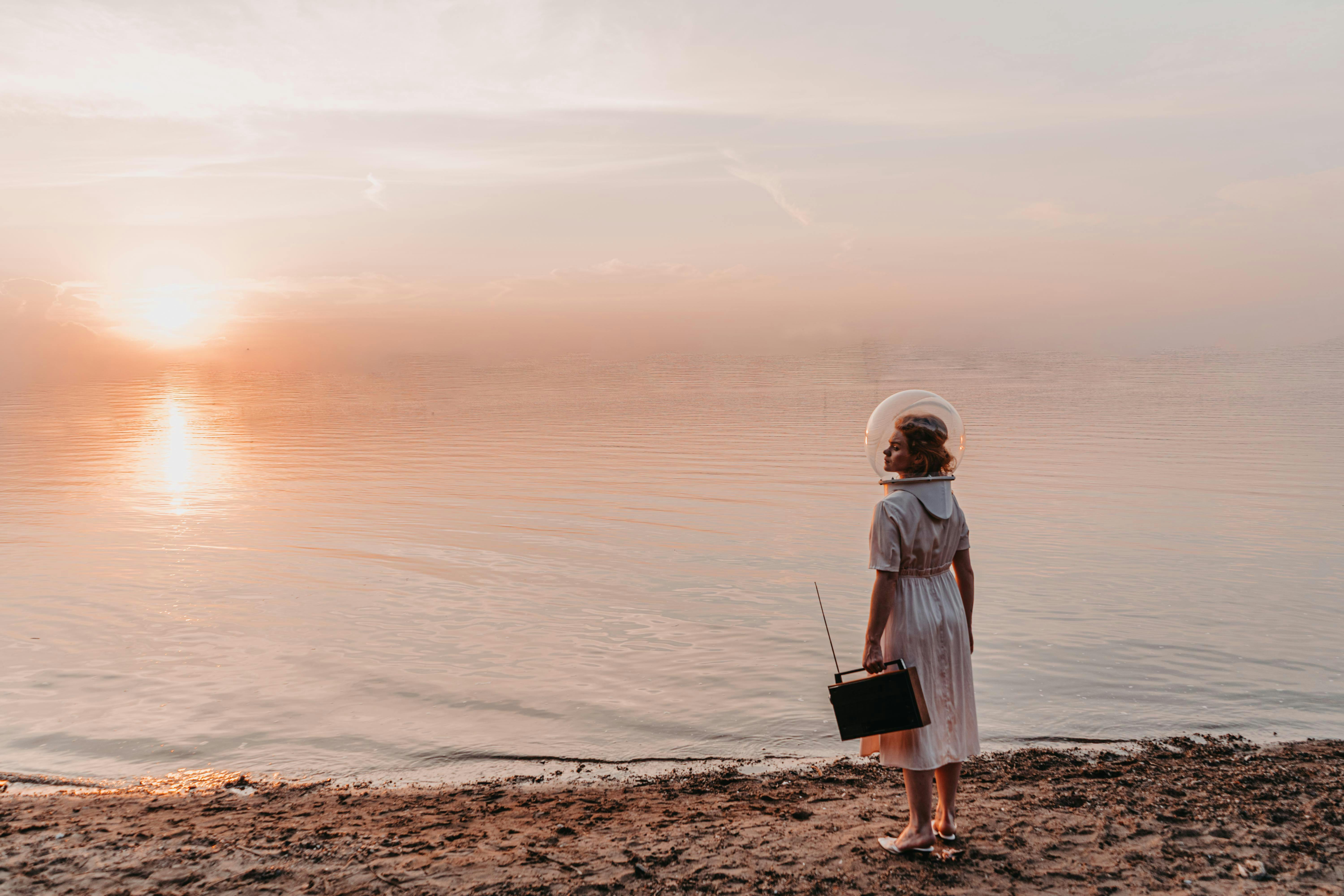 Person in retro attire with an astronaut helmet gazing at a serene beach sunset.