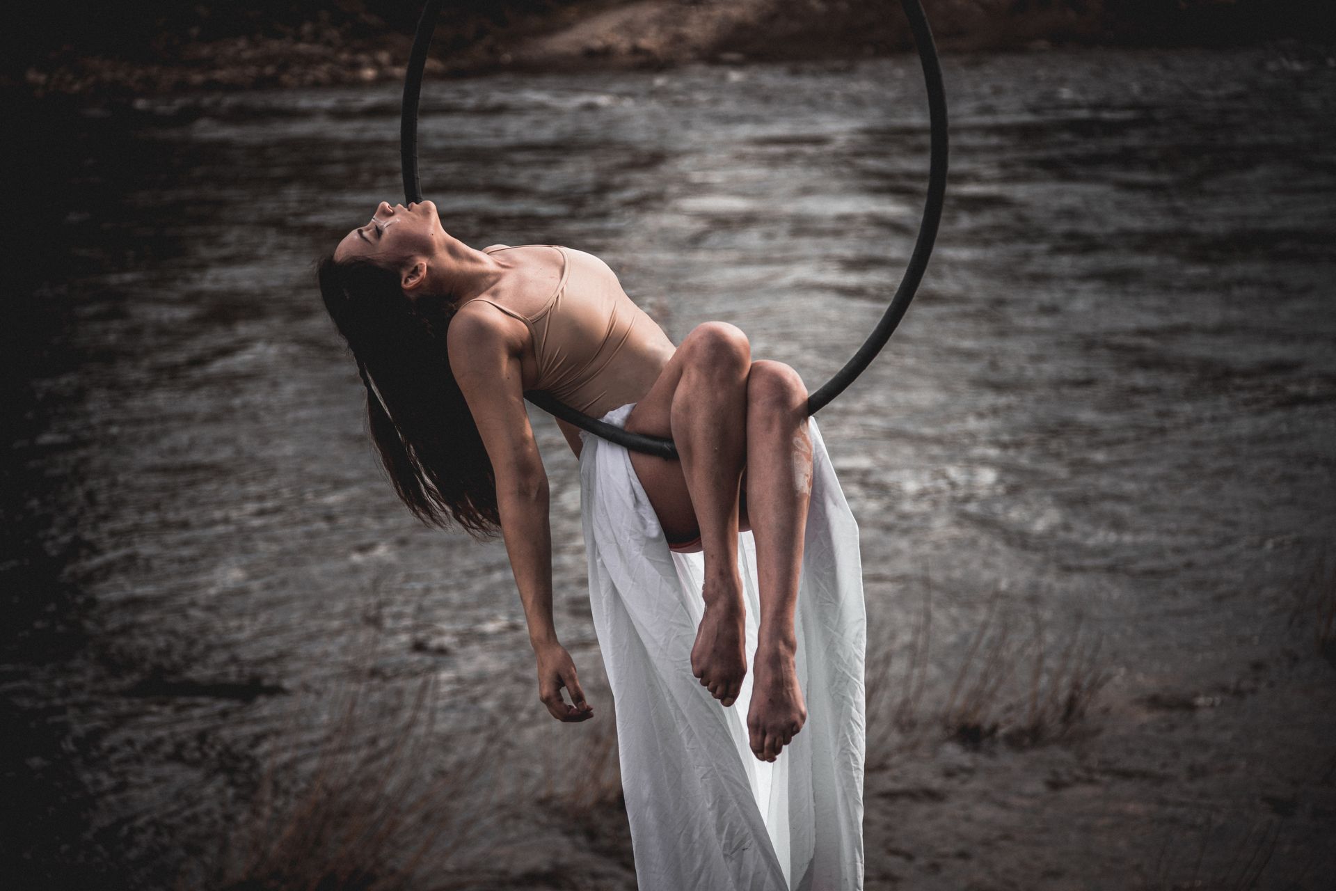 Woman on aerial hoop artfully poses by the water, embracing grace and strength.